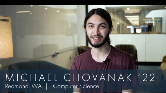 Michael Chovanak '22, Computer Science major from Redmond, WA smiles at the camera while sitting in a chair. There are glass walls and lounge chairs to the back of him.